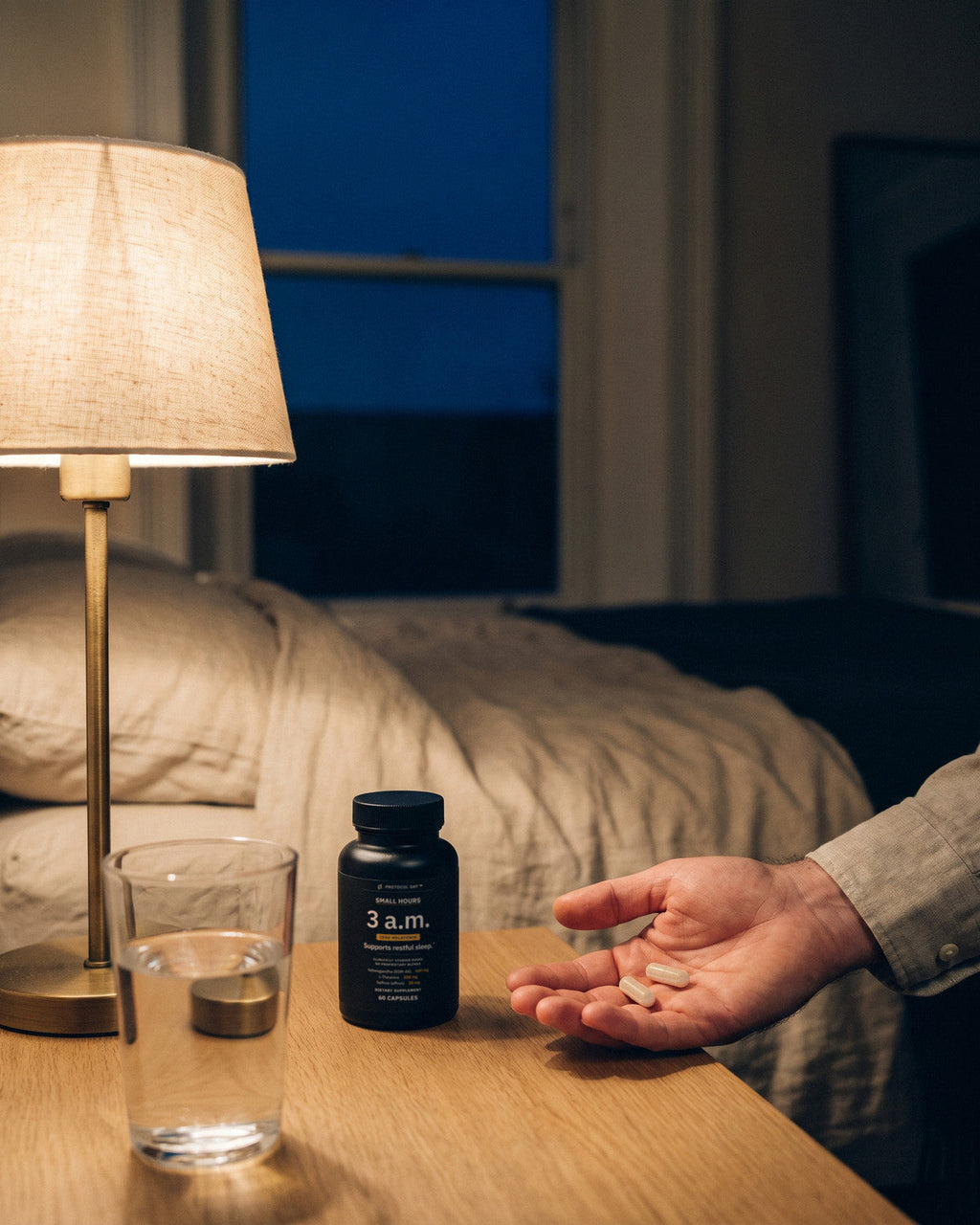 A hand holding two capsules by a bedside lamp, a glass of water and the Small Hours bottle on the table.